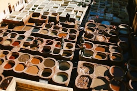 A traditional tannery with numerous circular pits filled with various colored dyes and tanned hides. Workers are seen moving between the pits, tending to the hides, and using long sticks. The scene is bathed in natural light, highlighting the rich and earthy tones of the dyes and leather.