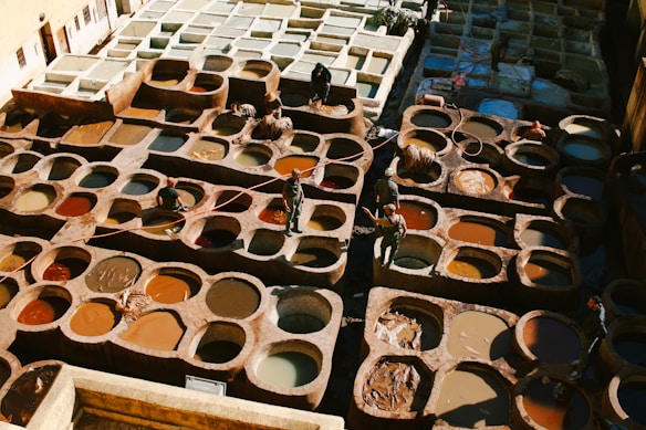 A traditional tannery with numerous circular pits filled with various colored dyes and tanned hides. Workers are seen moving between the pits, tending to the hides, and using long sticks. The scene is bathed in natural light, highlighting the rich and earthy tones of the dyes and leather.