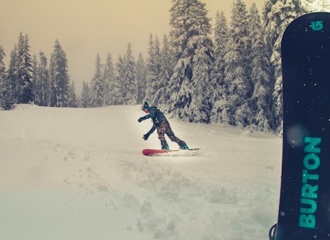 A person is snowboarding down a snowy slope surrounded by tall, snow-covered pine trees. The snowboarder is wearing a jacket, pants, and a beanie. The snowboard is partially visible on the right side with 'BURTON' branding in blue. The sky appears overcast, adding a muted tone to the scene.