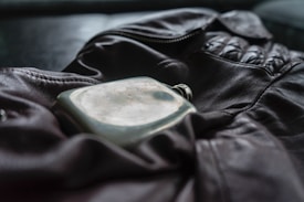 A leather jacket is laid out with a metal flask resting on top. The jacket features a prominent zipper and gathered sections at the shoulders, suggesting a sense of rugged style. The flask has a slightly worn appearance, reflecting a sense of nostalgia or adventure.