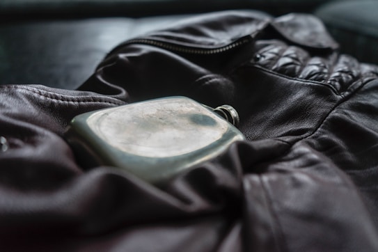 A leather jacket is laid out with a metal flask resting on top. The jacket features a prominent zipper and gathered sections at the shoulders, suggesting a sense of rugged style. The flask has a slightly worn appearance, reflecting a sense of nostalgia or adventure.