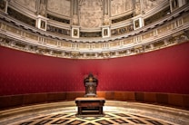 An ornate, historical room with a richly decorated throne in the center. The chair is elaborately carved, positioned against a backdrop of deep red walls that feature intricate patterns. The upper section of the room is adorned with detailed frescoes and classical sculptures. The flooring displays a complex black and white geometric pattern, adding to the overall opulence.
