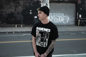 A young person is standing in an urban setting, wearing a black beanie and a Ramones T-shirt. The area has a gritty, industrial feel with a closed garage door and graffiti in the background. The person appears to have tattoos on their arm and is looking away from the camera, with a contemplative expression.