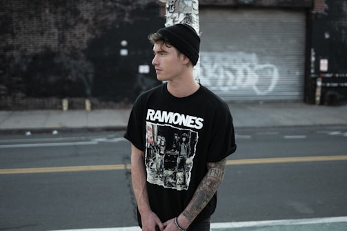 A young person is standing in an urban setting, wearing a black beanie and a Ramones T-shirt. The area has a gritty, industrial feel with a closed garage door and graffiti in the background. The person appears to have tattoos on their arm and is looking away from the camera, with a contemplative expression.