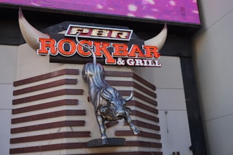 A large metallic bull sculpture is mounted on a wall with a prominent sign above it reading 'ROCKBAR & GRILL'. The sign features neon-like, bold letters, and the bull appears to be in a dynamic, charging pose. The background includes geometric wooden patterns, and there are large stylized horns framing the top part of the sign.