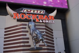 A large metallic bull sculpture is mounted on a wall with a prominent sign above it reading 'ROCKBAR & GRILL'. The sign features neon-like, bold letters, and the bull appears to be in a dynamic, charging pose. The background includes geometric wooden patterns, and there are large stylized horns framing the top part of the sign.