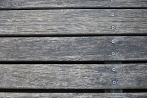 Wooden planks aligned horizontally, featuring a weathered texture with visible grain patterns. Several metal screws are evenly spaced along the center of each plank, securing them together.
