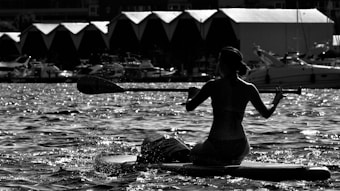 A silhouette of a person paddling on a kayak across a body of water with several boats docked in the background. The lighting creates a high contrast effect on the water's surface, highlighting splashes and ripples.