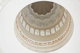 An intricately designed dome featuring delicate patterns and motifs. Arabic script adorns the circular band around the dome, and the architecture showcases both geometric and floral designs, with sections that allow natural light to filter through.