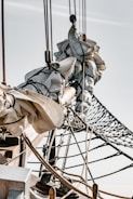 The image displays a close-up view of a ship's rigging, focusing on tightly coiled sailcloth, ropes, and a net against a clear sky. The wooden masts and lines are intricately detailed, showcasing the craftsmanship of nautical equipment.