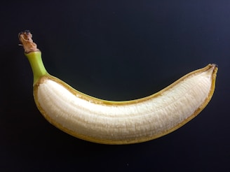 A banana with its peel partially removed, revealing the light yellow flesh inside, is set against a dark background. The fruit appears fresh and ripe.
