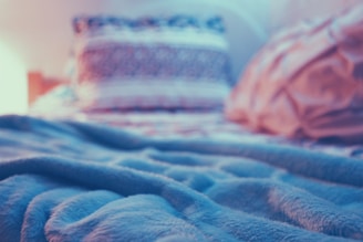 A cozy, soft blanket lies on a bed with two blurred pillows in the background. The lighting is warm, giving a serene and peaceful atmosphere.