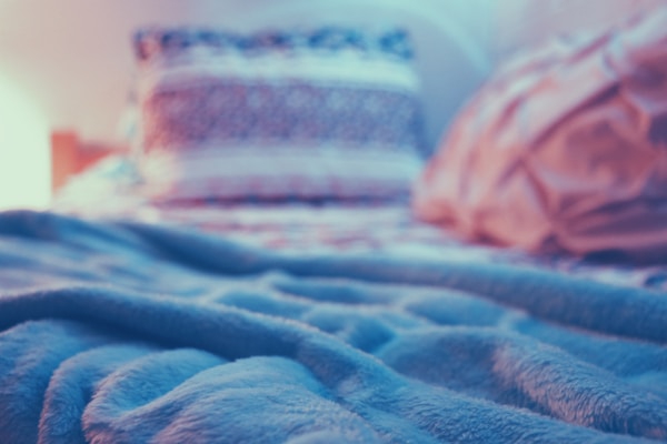 A cozy, soft blanket lies on a bed with two blurred pillows in the background. The lighting is warm, giving a serene and peaceful atmosphere.