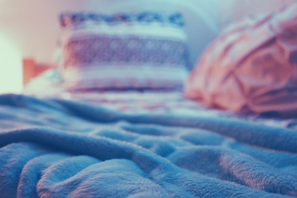 A cozy, soft blanket lies on a bed with two blurred pillows in the background. The lighting is warm, giving a serene and peaceful atmosphere.
