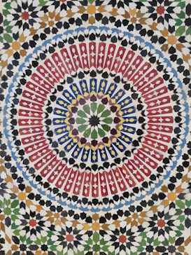 A vibrant and intricate geometric mosaic pattern featuring concentric circles with repeating shapes and an array of colors, including red, blue, green, yellow, and black. The design is symmetrical with a kaleidoscope effect, showcasing traditional Islamic art style with detailed craftsmanship.