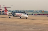 A SpiceJet propeller airplane is parked on the tarmac of an airport. The aircraft has a white body with red accents and the SpiceJet logo prominently displayed. Several airport vehicles and equipment can be seen in the background with a few people walking around. The sky is overcast, casting a muted light over the scene.