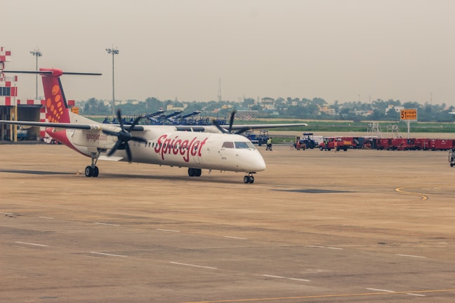 A SpiceJet propeller airplane is parked on the tarmac of an airport. The aircraft has a white body with red accents and the SpiceJet logo prominently displayed. Several airport vehicles and equipment can be seen in the background with a few people walking around. The sky is overcast, casting a muted light over the scene.