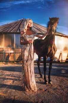 A woman in an elegant, sparkling gown stands next to a black horse, gently holding its reins. The background features a round wooden structure and a clear blue sky, with sunlight casting a warm glow.