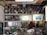A workshop with shelves filled with metal cylinders and various tools. A person wearing a hat is working at a cluttered bench, surrounded by equipment and metal parts. Two illuminated paintings are displayed on the wall, depicting nature scenes. The workspace has an industrial and artistic feel.