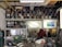 A workshop with shelves filled with metal cylinders and various tools. A person wearing a hat is working at a cluttered bench, surrounded by equipment and metal parts. Two illuminated paintings are displayed on the wall, depicting nature scenes. The workspace has an industrial and artistic feel.