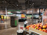 A grocery store produce section with neatly arranged vegetables and fruits. Shelves are filled with items like tomatoes, mushrooms, and colorful produce. Bright overhead lighting illuminates the area. Signs display prices and product information. People are seen walking and shopping.