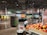 A grocery store produce section with neatly arranged vegetables and fruits. Shelves are filled with items like tomatoes, mushrooms, and colorful produce. Bright overhead lighting illuminates the area. Signs display prices and product information. People are seen walking and shopping.
