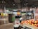A grocery store produce section with neatly arranged vegetables and fruits. Shelves are filled with items like tomatoes, mushrooms, and colorful produce. Bright overhead lighting illuminates the area. Signs display prices and product information. People are seen walking and shopping.