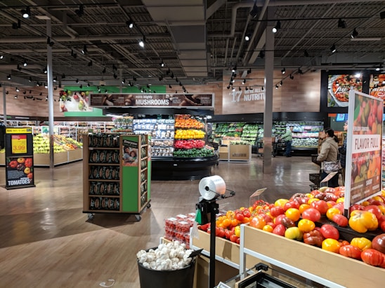 A grocery store produce section with neatly arranged vegetables and fruits. Shelves are filled with items like tomatoes, mushrooms, and colorful produce. Bright overhead lighting illuminates the area. Signs display prices and product information. People are seen walking and shopping.