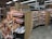 A supermarket aisle dedicated to pasta and sauce, featuring wooden shelving filled with jars of tomato sauce, packets of pasta, and various canned goods. The shelves are neatly organized, displaying a variety of brands and products. Decorative items like wheat bundles and artificial vegetables are placed around the display.