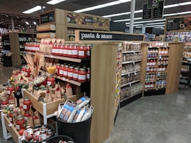 A supermarket aisle dedicated to pasta and sauce, featuring wooden shelving filled with jars of tomato sauce, packets of pasta, and various canned goods. The shelves are neatly organized, displaying a variety of brands and products. Decorative items like wheat bundles and artificial vegetables are placed around the display.