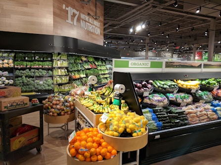 A well-organized produce section within a grocery store featuring a wide variety of fresh fruits and vegetables. The display includes items like oranges, bananas, lemons, and an array of leafy greens. The section is labeled 'Organic' and the signage emphasizes nutrition. The lighting is bright and the overall setup is clean and orderly.