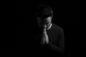 A person in a dark environment with a focused expression and hands held together in a praying or contemplative gesture. The figure is positioned centrally, and the lighting creates a dramatic shadow effect over the face and body.