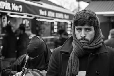 A man with facial hair and a scarf around his neck stands in the foreground, looking directly at the camera with a serious expression. He wears a coat, and the background features several people blurred out, including a person in a headscarf. The scene appears to be a bustling street or marketplace.