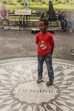 A child wearing a red shirt with a Mario graphic is standing on a mosaic circle with the word 'IMAGINE' inscribed on it. In the background, there are people sitting on a bench, with one playing a guitar. A sign on the bench reads 'RIP Aretha Queen of Soul'. The surrounding area is a park with green grass and trees.