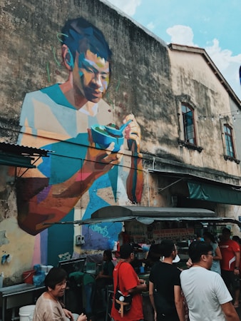 A vibrant mural depicting a person enjoying a bowl of food is painted on an old building wall. Below the mural, a small group of people gather around a street food stall. The atmosphere is lively and social, with several people in casual clothing interacting.