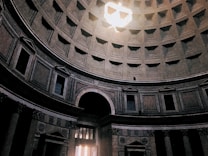 The image depicts the interior of a grand, ancient dome with intricate architectural details. Sunlight beams through an oculus at the top, illuminating the space with a dramatic ray of light. The walls are lined with recessed panels and decorative pilasters, creating a sense of depth and historical grandeur.