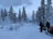 A snowy landscape with tall evergreen trees covered in snow. Two people wearing winter gear stand near snowmobiles, ready for a winter adventure. The sky is overcast, with hints of pink and blue colors peeking through the clouds.