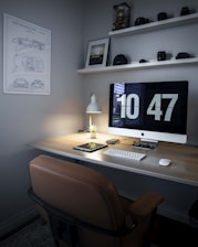 A neatly organized home office desk setup featuring a large monitor displaying the time 10:47, a sleek table lamp, a small potted plant, a wireless keyboard and mouse, and a tablet. The wall is decorated with a poster showing car schematics and shelves holding cameras and lenses.