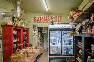 A rustic store interior with a sign reading 'Barnaroo' above a commercial refrigerator filled with various products. The room is filled with shelves carrying baskets, canned goods, jars, and decorative items. A red cabinet on the left holds a scale and other products, while hats and other items are placed on shelves.