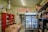 A rustic store interior with a sign reading 'Barnaroo' above a commercial refrigerator filled with various products. The room is filled with shelves carrying baskets, canned goods, jars, and decorative items. A red cabinet on the left holds a scale and other products, while hats and other items are placed on shelves.