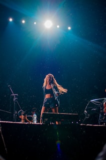 A performer is energetically dancing on a stage under bright stage lights. The scene includes several spotlights above, illuminating the stage, and there appears to be mist or rain adding to the dynamic atmosphere. A microphone and a water bottle are visible, suggesting a live performance setting.