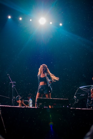 A performer is energetically dancing on a stage under bright stage lights. The scene includes several spotlights above, illuminating the stage, and there appears to be mist or rain adding to the dynamic atmosphere. A microphone and a water bottle are visible, suggesting a live performance setting.