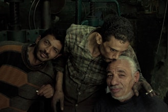Three men are gathered closely together in a dimly lit setting. The man on the left is leaning toward the camera with a neutral expression, wearing a striped sweater. The man in the center is bowing his head slightly, expressing affection by planting a gentle kiss on the forehead of the man sitting at the front. The man receiving the kiss is smiling gently, displaying a sense of warmth and camaraderie. The background is dark and industrial, possibly in a workshop or factory.