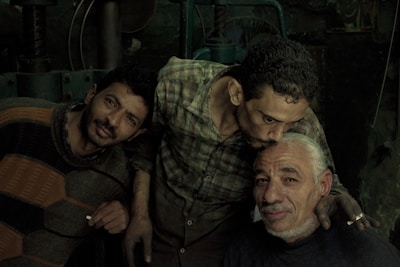 Three men are gathered closely together in a dimly lit setting. The man on the left is leaning toward the camera with a neutral expression, wearing a striped sweater. The man in the center is bowing his head slightly, expressing affection by planting a gentle kiss on the forehead of the man sitting at the front. The man receiving the kiss is smiling gently, displaying a sense of warmth and camaraderie. The background is dark and industrial, possibly in a workshop or factory.