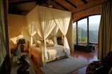 A cozy bedroom with a canopy bed draped with white curtains, surrounded by warm, earthy tones. The room features a mix of rustic and elegant décor, including vases and a lamp. Large glass doors open to a stunning mountain view, allowing natural light to flood the space.
