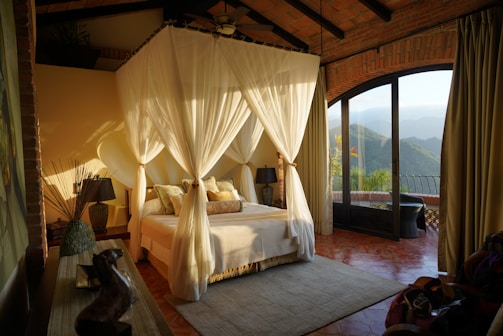 A cozy bedroom with a canopy bed draped with white curtains, surrounded by warm, earthy tones. The room features a mix of rustic and elegant décor, including vases and a lamp. Large glass doors open to a stunning mountain view, allowing natural light to flood the space.