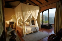 A cozy bedroom with a canopy bed draped with white curtains, surrounded by warm, earthy tones. The room features a mix of rustic and elegant d&eacute;cor, including vases and a lamp. Large glass doors open to a stunning mountain view, allowing natural light to flood the space.