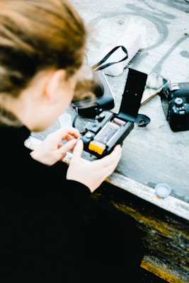 A person is seen from behind, focusing intently on operating a film camera. The person is adjusting the camera's settings or loading film. Nearby on the table lie a digital camera, a smartphone, and a few other items like a camera lens and a strap.