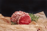 A raw piece of marbled beef is resting on crumpled brown parchment paper. Fresh rosemary sprigs and a few scattered red peppercorns add to the presentation. The meat is vibrant red with intricate patterns of white fat.
