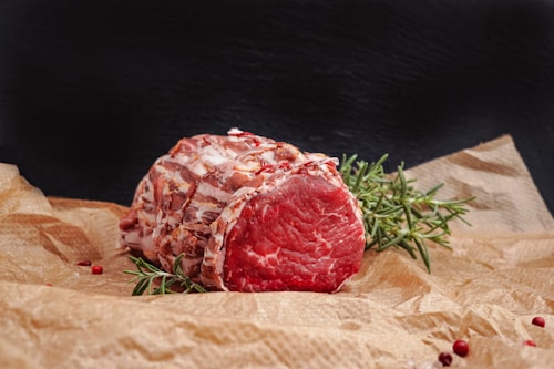 A raw piece of marbled beef is resting on crumpled brown parchment paper. Fresh rosemary sprigs and a few scattered red peppercorns add to the presentation. The meat is vibrant red with intricate patterns of white fat.